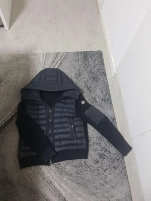 Svart quiltad pufferjacka med huva - Snygg svart pufferjacka med quiltad front och huva. Jackan har stickade ärmar och muddar, dragkedja framtill samt fickor med dragkedja. En patchdetalj syns på ena ärmen. Perfekt för kyligare dagar och ger en sportig look.