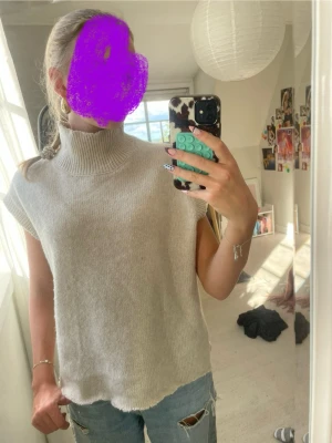 Beige stickad ärmlös polotröja - Säljer en beige stickad ärmlös tröja med hög polokrage. Tröjan har en avslappnad passform och är perfekt att styla över en långärmad topp eller t-shirt. Materialet ser mjukt och mysigt ut, och färgen är lätt att matcha med det mesta.