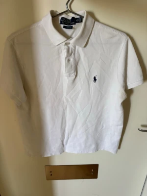 Vit pikétröja från Polo Ralph Lauren - Klassisk vit pikétröja från Polo Ralph Lauren med korta ärmar och krage. Tröjan har knappar framtill och den ikoniska broderade loggan på bröstet. Perfekt till jeans eller shorts för en clean och stilren look. Storlek M, Custom Fit