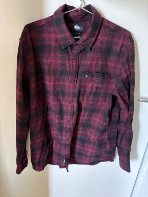 Röd och svart rutig skjorta - Säljer en röd och svart rutig skjorta från Quiksilver med klassisk krage, knappstängning och en bröstficka. Skjortan har långa ärmar och är tillverkad i ett mjukt tyg, perfekt för en avslappnad stil.