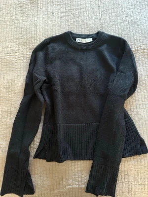 Svart stickad tröja från ZARA - Snygg svart stickad tröja från ZARA med rund halsringning och ribbade detaljer vid ärmslut och nederkant. Tröjan har långa ärmar och slitsar på sidorna för en modern look. Perfekt till jeans eller kjol. Slitsar vid ärmarna.