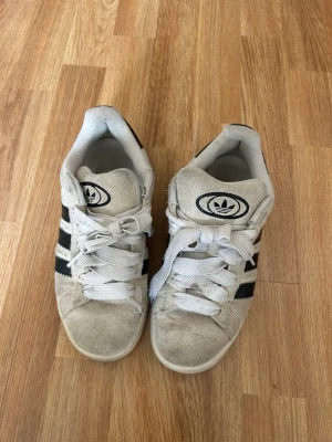 Adidas campus  - Säljer ett par beige sneakers från Adidas med klassiska svarta ränder på sidorna och Adidas-logga på plösen. Skorna har vita snören och en gummisula i brunt. Ovandelen är i mocka och modellen är låg.