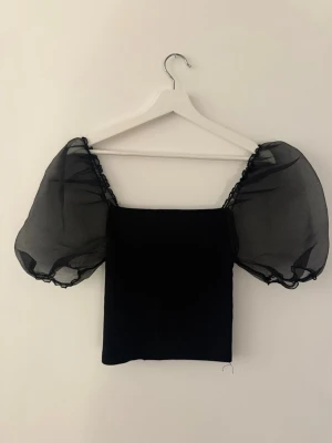 Svart topp med puffärmar - Trendig svart topp med stora puffärmar i transparent material. Toppen har en rak, croppad passform och fyrkantig urringning. Perfekt för dig som vill sticka ut med en cool och modern look.