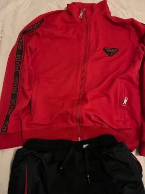 Tracksuit från Prada - Snygga svarta mjukisbyxor från Prada med röda detaljer och dragsko i midjan. Byxorna har fickor med dragkedja och en diskret Prada-logga på benet. Perfekta för en chill och trendig look. Med koftan.