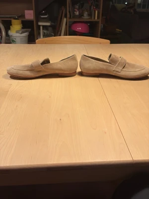 Beige mockaloafers med låg sula - Säljer ett par stilrena loafers i beige mocka med klassisk penny strap och låg, ljusbrun sula. Skorna har rundad tå och enkel design som passar till många olika outfits. Perfekta för dig som gillar en clean och avslappnad look.