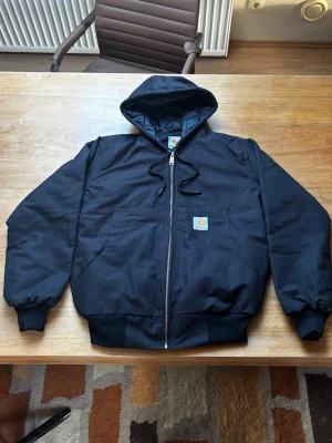 Carhartt jacka - Säljer en svart jacka från Carhartt med dragkedja framtill, huva med dragsko och två stora fickor på magen. Klassisk Carhartt-logga på bröstet. Ribbstickade muddar vid ärmslut och nederkant. Perfekt för en chill och street-inspirerad look.