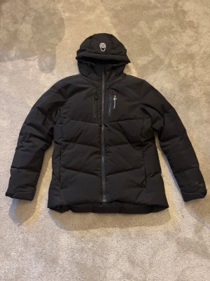 Svart dunjacka med huva - Snygg svart dunjacka med huva och dragkedja framtill. Jackan har en bröstficka med dragkedja och diskret logga på bröstet. Quiltad design och varm vaddering, perfekt för kalla vinterdagar. Jackan har en modern passform och är enkel att matcha.