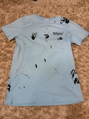 Ljusblå Off-White t-shirt med tryck - Säljer en ljusblå t-shirt från Off-White med svart grafiskt tryck och logga framtill samt stort ikoniskt tryck på ryggen. T-shirten har korta ärmar och är gjord i mjuk bomull. Perfekt för dig som gillar streetwear och vill sticka ut.