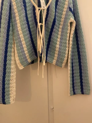 Randig stickad cardigan med knytband - Säljer en stickad cardigan med blå, ljusblå, vit och beigea ränder. Tröjan har långa ärmar, v-ringning och knytband framtill. Materialet är virkat och ger en luftig känsla. Perfekt för lager på lager och en cool retrostil.