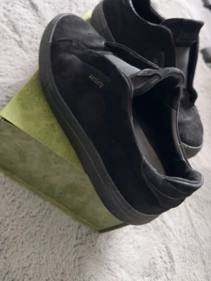 Svarta sneakers från Axel Arigato - Snygga svarta sneakers från Axel Arigato i mocka med diskret logga på sidan. Skorna har en låg profil och elastiskt band över vristen istället för klassiska snören. Perfekta för en clean och stilren look.
