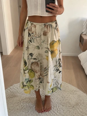 Långkjol med blommigt mönster - Luftig långkjol i bomull med elastisk midja och blommigt mönster i grönt, gult, brunt och beige på vit botten. Kjolen har en avslappnad passform och är perfekt för en somrig look.