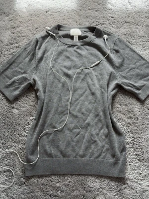 Grå basic stickad tshirt - En enkel grå stickad tröja med rund halsringning och korta ärmar. Tröjan har ribbade muddar vid ärmslut och nederkant, vilket ger en clean och stilren look. Perfekt att matcha med jeans eller kjol för en avslappnad stil.