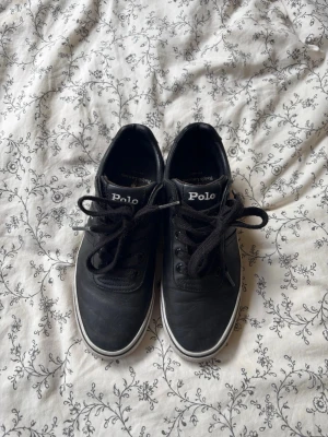 Svarta Polo Ralph Lauren sneakers - Snygga svarta sneakers från Polo Ralph Lauren med vita sulor och klassisk logga på sidan. Står storlek 38 på dem men de är lite små i storleken så skulle säga 37-37.5