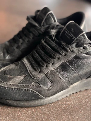 Valentino rockrunners - Snygga svarta sneakers från Valentino med diskret camomönster och detaljer i mocka och läder. Skorna har svarta skosnören och en klassisk låg siluett. Perfekta för dig som gillar stilrena och exklusiva sneakers.