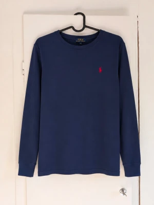 Ralph Lauren tröja - Säljer en klassisk vintage långärmad tröja från Polo Ralph Lauren i mörkblått med den ikoniska röda loggan på bröstet. En stilren och tidlös modell som passar perfekt året runt och är enkel att matcha med både jeans och chinos. Tröjan är i storlek L (14-16) och har en bekväm passform med mjukt material och minimalistisk design. Den här äldre Polo-modellen har den där genuina vintagekänslan som blivit allt mer eftertraktad. Självklart äkta✅