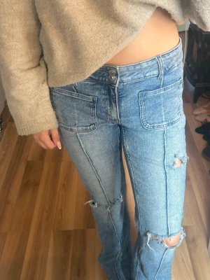 Blå slitna jeans med stora fickor - Säljer ett par ljusblå jeans med raka ben, stora framfickor och slitningar på knäna. Jeansen har mid waist och synliga sömmar som ger en cool vintagekänsla. Perfekta för dig som gillar en Sthlm stil och trendig stil.
