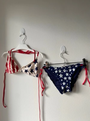 Bikini med stjärnor och ränder - Snygg bikini med trekantsöverdel och knytband i rött. Överdelen har ena kupan med röda och vita ränder, andra med blå stjärnor på vit botten. Underdelen är marinblå med vita stjärnor och röda knytband i sidorna. Perfekt för sommaren och strandhäng! Överdelen har öppning på sidorna ifall man vill lägga till inlägg.