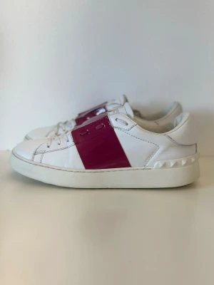 Vita valentino sneakers med lila band - Snygga vita sneakers med ett glansigt vinrött band över snörningen och coola nitar på hälen. Skorna har en klassisk låg profil och är tillverkade i läder med vita snören och sula. Perfekta för dig som vill sticka ut lite extra.