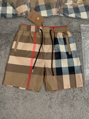 Burberry shorts - Säljer ett par klassiska Burberry shorts i den ikoniska rutiga designen med beige, svart, röd och vit färg. Shortsen har elastisk midja med svart snörning och sidofickor. Perfekta för sommaren och riktigt snygga till streetwear-stilen.