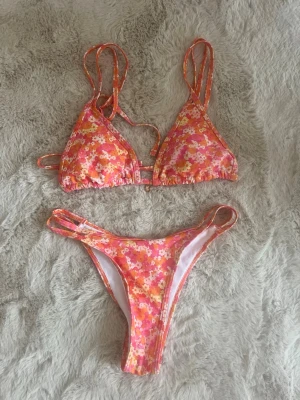 Blommig bikini - Snygg bikini med blommigt mönster i rosa, orange, gult och vitt. Överdelen har trekantskupor, dubbla axelband och en liten guldfärgad snäcka som detalj framtill. Underdelen har smala sidoband och matchar toppen perfekt, superfin och aldrig använd storlek S