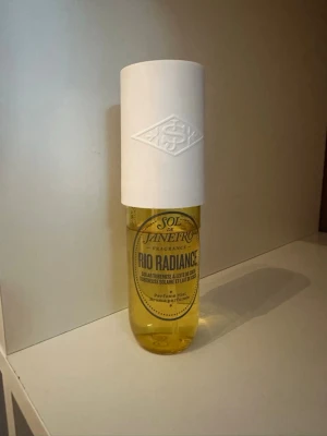 Sol de Janeiro Rio Radiance parfym - Sol de Janeiro Rio Radiance är en parfym i en genomskinlig gul flaska med en stor vit cylinderformad kork. Flaskan har en modern design och etiketten är dekorerad med svart text. Doften är inspirerad av brasiliansk sol och strand.