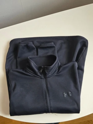 Svart zip-tröja från Under Armour - Säljer en svart zip-tröja från Under Armour med hög krage och dragkedja framtill. Tröjan har en diskret logga på bröstet och är tillverkad i ett mjukt, stretchigt material som passar perfekt till träning eller chill. Enkel och stilren design.