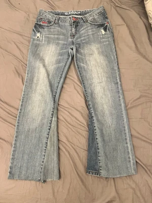 Ljusblå bootcut jeans från Guess - Säljer ett par utsydda ljusblå bootcut jeans från Guess med slitningar framtill och broderad bakficka. Jeansen har låg midja, röda detaljer och råa kanter vid bensluten. Perfekta för en avslappnad och trendig look. (Skriv till mig för mått💋)