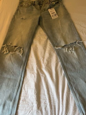 Ljusblå slitna jeans med hål Zara  - Säljer ett par ljusblå jeans med trendiga slitningar och stora hål vid knäna. Jeansen har rak passform och klassisk femficksdesign. Perfekta för en avslappnad och cool stil. Materialet är denim i bomull. Låg midja, säljer för att dom har för stora.