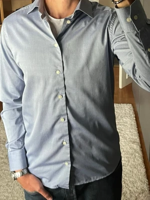 Eton skjorta i ljusblå - stl 40 (M) - Slim fit i 100% bomull med diskret vävd prickstruktur i tyget. Cutaway-krage, funkar både till kostym och till jeans. Personen på bilden är 189cm, väger 78kg.  Använd men i mycket fint skick, inga hål eller fläckar.  Rök- och djurfritt hem. Skriv gärna om du vill ha fler bilder eller undrar något!