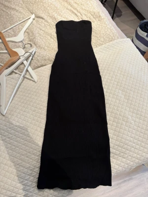 Svart ribbad axelbandslös klänning - Lång svart klänning i ribbat material med axelbandslös design. Klänningen har en tight passform och är perfekt för dig som gillar en stilren och enkel look. Passar till många olika tillfällen och är lätt att styla med accessoarer.