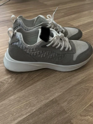 Gråvita sneakers  - Snygga sneakers från  i grått och vitt med meshdetaljer och mockaliknande partier. Skorna har vita snören, tjock vit sula och diskret logga på plösen. Perfekta för dig som gillar stilrena och trendiga sneakers.
