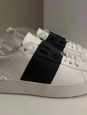 Vita sneakers med svart detalj Valentino - Stilrena vita sneakers från Valentino med en bred svart läderdetalj över sidan. Skorna har vita snören, låg profil och en mönstrad sula med Valentino-logga undertill. Tillverkade i slätt läder för en lyxig känsla.