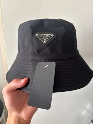 Svart bucket hat från Prada - Snygg svart bucket hat från Prada med det klassiska trekantiga metallogot på framsidan. Hatten är tillverkad i ett glansigt, slitstarkt material och har en bred, nedåtlutande brätte. Perfekt accessoar för en trendig och avslappnad stil.