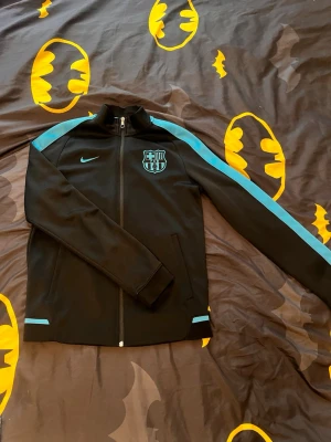 Svart Nike FC Barcelona träningsjacka - Snygg svart träningsjacka från Nike med FC Barcelona-logga på bröstet och turkosa detaljer längs ärmarna och vid muddar. Jackan har hel dragkedja framtill, ståkrage och två sidofickor. Perfekt för dig som älskar fotboll och vill ha en sportig look.