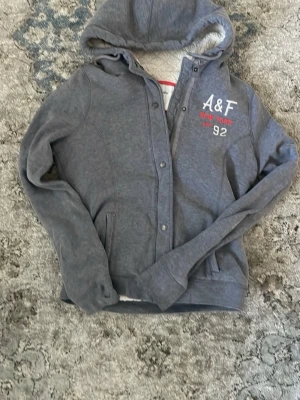 Grå hoodie från Abercrombie & Fitch - Mysig grå hoodie från Abercrombie & Fitch med broderad text 'A&F New York EST 92' i vitt och rött på bröstet. Tröjan har dragkedja, knappar, två fickor fram och en fodrad huva med mjukt material. Perfekt för chill dagar.