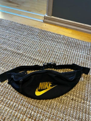 Svart midjeväska från Nike - Snygg svart midjeväska från Nike med guldfärgad logga framtill. Väskan har justerbart band och spänne, samt dragkedja upptill. Perfekt för att bära det viktigaste när du är på språng. Tillverkad i slitstarkt tygmaterial.