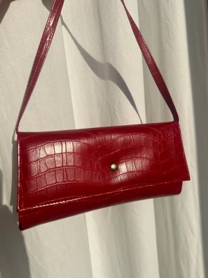 Vintage röd kuvertväska i krokopräglat skinn - Snygg röd kuvertväska i krokopräglat konstläder med axelrem och guldfärgad knappstängning. Väskan har ett stilrent, avlångt format och insida i matchande rött tyg. Perfekt för hösten!! Med avtagbara band så man kan anväds den som en pouch🤩❤️
