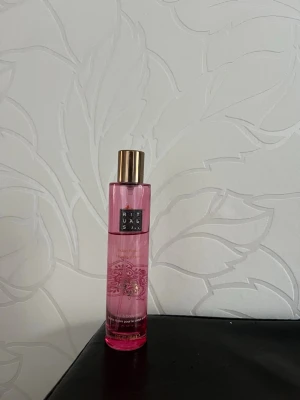 Rituals rosa parfymflaska - Säljer en snygg parfym från Rituals i en cylinderformad rosa glasflaska med guldfärgat lock. Flaskan har dekorativa mönster och text i vitt och guld. Perfekt för dig som gillar fräscha och blommiga dofter.