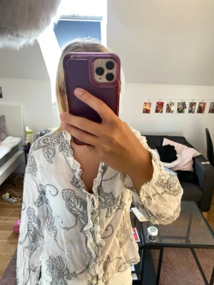 Vit blus med volang och svart mönster - Säljer en vit blus med svart paisley-liknande mönster. Skjortan har volangdetaljer längs knappraden och är långärmad. Materialet är lätt och luftigt, perfekt för vår och sommar. Knappar framtill och en feminin touch med volanger.