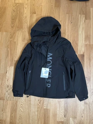 Svart vindjacka från Moncler - Svart vindjacka från Moncler med stor logotyp längs dragkedjan. Jackan har huva, två dragkedjeförsedda fickor och justerbar nederkant. Materialet är lätt och vattenavvisande, perfekt för blåsiga dagar. Snygg och stilren design med sportig känsla.