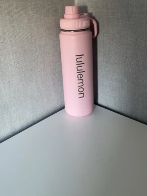 Lululemon vattenflaska 710 ml - Snygg rosa vattenflaska från Lululemon med skruvlock och praktisk bärögla. Flaskan är tillverkad i tålig plast och har en stilren design med Lululemon-loggan tryckt längs sidan. Perfekt att ha med till gymmet, skolan eller på språng.