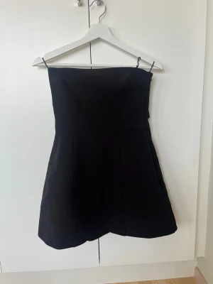 Svart axelbandslös kort klänning från zara  - Snygg svart axelbandslös kort klänning med stor dekorativ rosett baktill. Klänningen har en enkel, elegant siluett och är perfekt för dig som vill sticka ut. Tillverkad i ett slätt material som ger en stilren look. Helt ny från zara! 