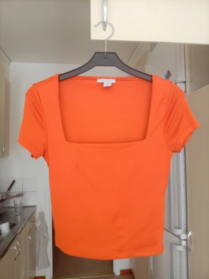 Orange topp med fyrkantig ringning Amisu - Säljer en orange topp från Amisu i storlek S. Toppen har en snygg fyrkantig ringning och korta ärmar. Materialet är mjukt och stretchigt, perfekt för en trendig och bekväm look. Passar till många olika stilar och outfits.