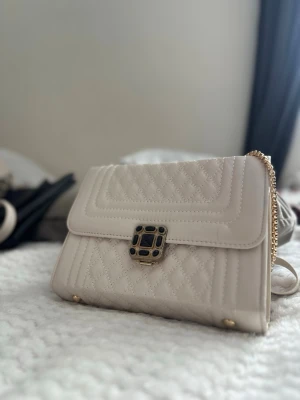 Beige quiltad handväska med kedja - Stilren beige handväska i quiltat mönster med guldfärgad kedjerem och dekorativt spänne framtill. Väskan har en rektangulär form och detaljer i guld som ger en lyxig känsla. Perfekt accessoar för att lyfta din outfit.
