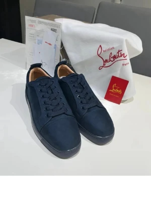 Marinblå sneakers från Louboutin - Snygga marinblå sneakers från Christian Louboutin med klassisk röd sula och lågt skaft. Skorna har ovandel i mocka, matchande marinblå snören och diskreta sömmar. Insidan är ljusbeige och sulan har Louboutin-logga i rött. Perfekta för dig som gillar exklusiv streetstyle.