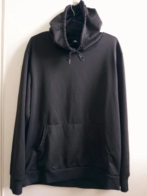 Svart hoodie med ficka och huva - En stilren svart hoodie med stor känguruficka framtill och justerbar huva med dragsko. Tillverkad i mjukt bomullsmaterial och har ribbade muddar vid ärmslut och nederkant. Perfekt för en avslappnad och enkel look.