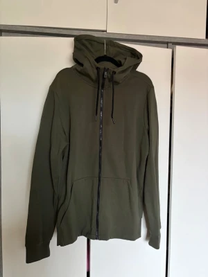 Olivgrön hoodie från SOC - Säljer en olivgrön hoodie från SOC i storlek L. Hoodien har dragkedja framtill, två fickor och justerbar huva med snören. 