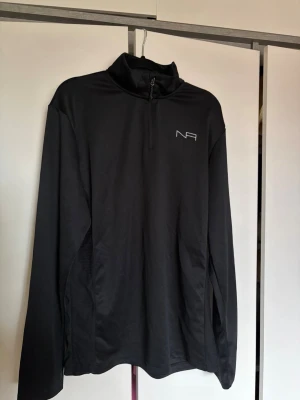 Svart träningströja - Snygg svart långärmad träningströja från New Apparel med halv dragkedja och hög krage. Tillverkad i ett lätt och ventilerande material, perfekt för löpning eller gym. 