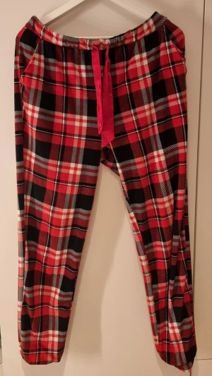 Rödrutiga pyjamasbyxor i velour med snöre - Mysiga pyjamasbyxor i rött, svart och vitt rutmönster. Byxorna har resår i midjan och ett rött knytband framtill. Tillverkade i velour, perfekta för chillkvällar hemma. Klassisk design med sidofickor.