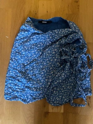 Blå blommig omlottkjol - Söt blå omlottkjol från BikBok i storlek XS med vitt blommönster och knytband i sidan. Kjolen har en kort längd och är perfekt för varma dagar. Materialet känns lätt och luftigt, vilket gör den skön att bära.🌸 Använd fåtal gånger